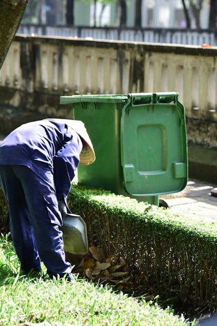 A gardener wearing a wide-brimmed hat and a dark blue jacket is crouched down, placing garden waste into a large green wheeled bin on a paved pathway. The bin's lid is open, revealing the collection o